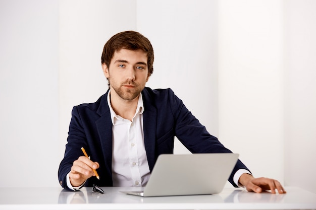 Free photo successful businessman sit at his office, working on project with laptop, hold pencil and look