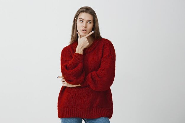 Successful businesswoman deciding how to escape awkward situation. Shot of serious gorgeous european woman in red loose sweater, holding hand above chin and thinking, being intense over gray wall