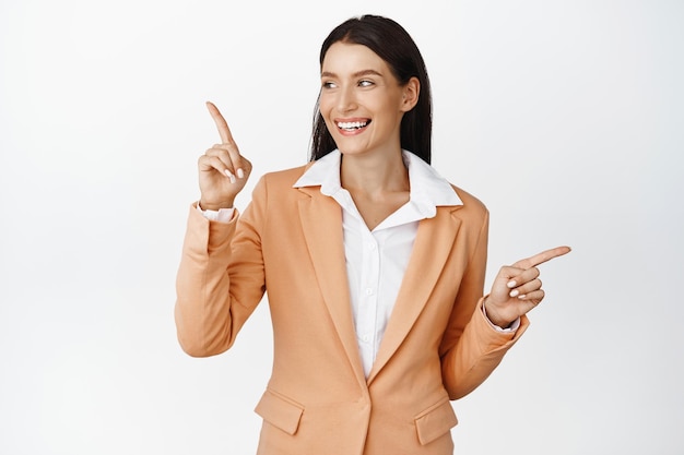 Successful corporate woman in suit smiling pointing fingers sideways looking left at logo company advertisement standing against white background