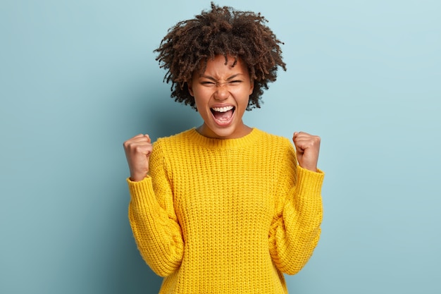 Successful dark skinned female student happy to get scholarship, clenches fists, accomplishes goal