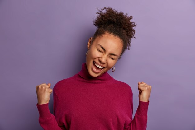 Successful euphoric African American woman celebrates amazing news, lucky to win lot of money, triumphs as dream come true, tilts head, dressed in casual turtleneck, isolated on purple wall