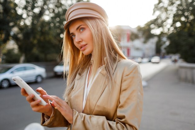 Successful fashionable european woman in elegant casual outfit posing mobyle phone outdoor. Sunset colors.