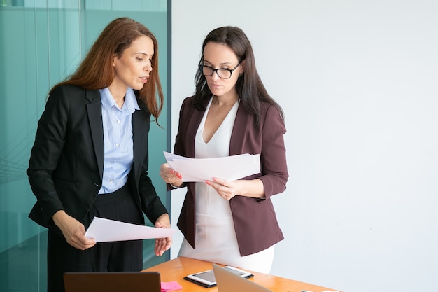 Successful female colleagues holding documents, reading report and standing in conference room together