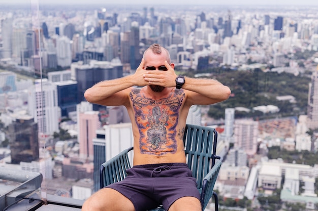 Free Photo successful handsome european bearded brutal tattooed strong man topless with watch sits on chair on high floor with amazing city view