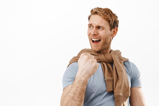 Successful handsome redhead man clench fist say yes and triumphing celebrate victory achieve goal looking left with pleased and satisfied face cheering standing over white background