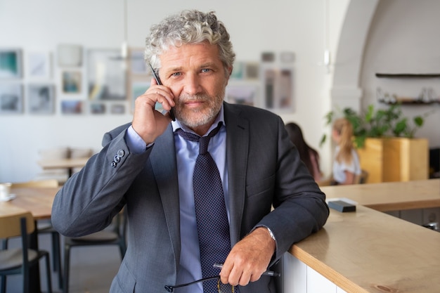 Successful mature businessman talking on mobile phone, standing at co-working, leaning on desk, looking at camera a