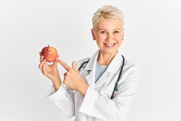 Successful middle aged woman physician wearing medical unifrom smiling at camera and pointing fore finger at ripe red apple which in good for gut health and promotes weight loss. Healthcare and diet