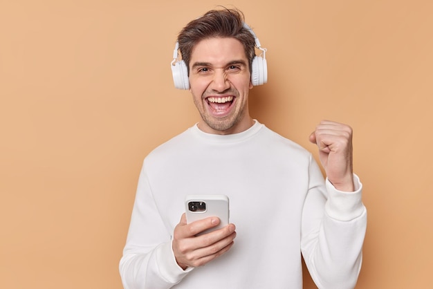 Free Photo successful positive man with dark hair clenches teeth rejoices good news wears casual white jumper uses smartphone for playing games online uses wireless headphones isolated over beige wall.