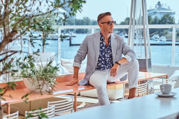 Free photo successful stylish man dressed in modern elegant clothes sitting on a table at outdoor cafe against the background of city wharf.