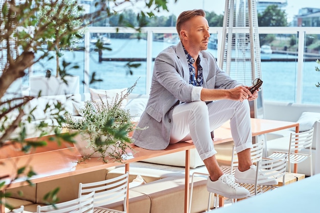 Free photo successful stylish man dressed in modern elegant clothes sitting on a table at outdoor cafe against the background of city wharf.