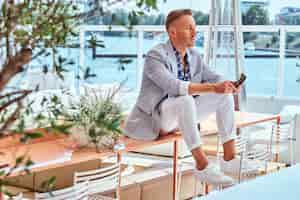 Free photo successful stylish man dressed in modern elegant clothes sitting on a table at outdoor cafe against the background of city wharf.