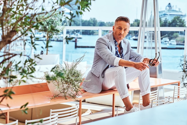 Successful stylish man dressed in modern elegant clothes sitting on a table at outdoor cafe against the background of city wharf.