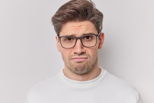 Sulking dissatisfied adult European man looks sadly at camera frowns angrily wears transparent glasses and casual jumper isolated over white background stands upset complains about something