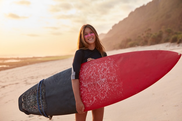 Summer activities concept. Satisfied beautiful young woman dressed in swimsuit, carries long board, has holidays abroad in resort country