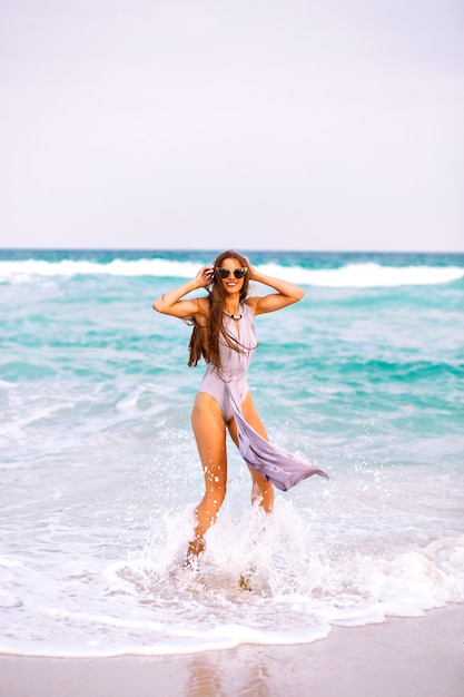 Free photo summer beach portrait of pretty brunette woman with sportive perfect tanned body and long brunette hairs, wearing trendy glamour elegant swimsuit, model relax near ocean, spin and having fun, freedom.