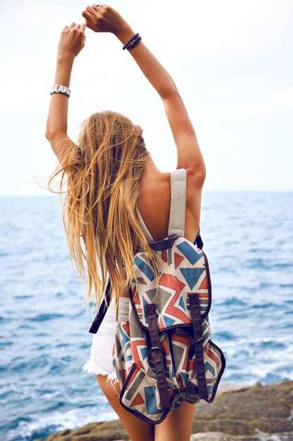 Summer fashion portrait go young woman traveling with backpack in summer time, posing near ocean at rainy day, wind sad mood.