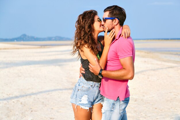 Summer fashion portrait of young pretty stylish hipsters couple in love hugs and posing at amazing island beach, having fun alone, wearing bright casual clothes and sunglasses.