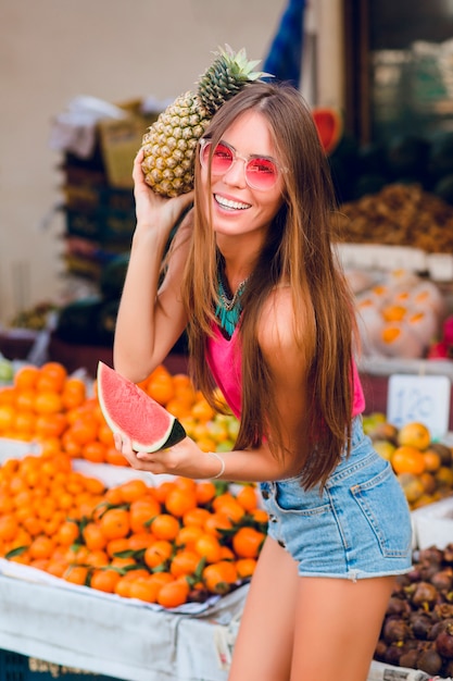 Free photo summer fashionable girl is posing with ananas and slice of watermelon on tropical fruits market