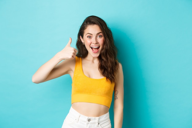 Summer holidays and emotions concept. Cheerful young woman saying yes, praising good choice, nice work, showing thumbs up and looking excited, standing over blue background