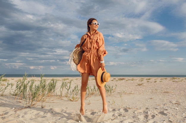 Summer image of beautiful brunette female in trendy linen dress jumping and fooling around , holding straw bag. Pretty slim girl enjoying weekends near ocean. Full length.