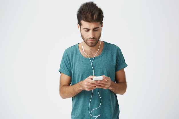 Free Photo sun-tanned fellow with stylish haircut messaging with friends on white wall. young man wearing earphones, looking  while answering to his girlfriend's message using wi-fi