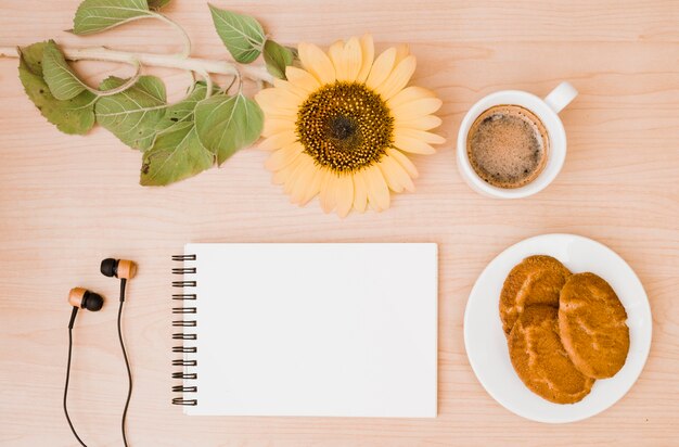 Sunflower twig; coffee cup; earphone; cookies and blank spiral notepad on wooden desk