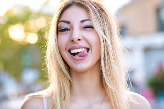 Sunny close up portrait of pretty blonde woman winking and showing tongue, playful positive mood, pastel sunny colors.