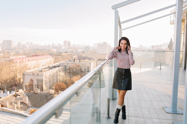 Sunny morning in big city on terrace of young brunette fashionable woman talking on phone and smiling. Attractive model woman, chilling, relaxing time, happiness.