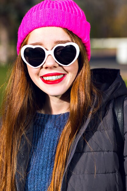 Sunny spring portrait of happy cheerful smiling ginger woman posing at the park, enjoy sunny day, bright punk hipster hat, hearted sunglasses, red lips, warm parka, positive mood.