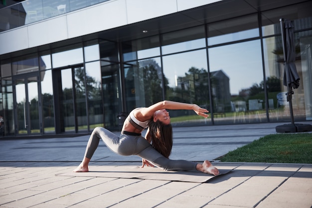 Sunny summer morning. Young athletic woman doing handstand on city park street among modern urban buildings. Exercise outdoors healthy lifestyle