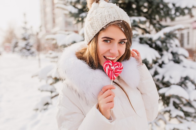 Free photo sunny winter morning on street of charming young woman licking pink lollypop. happy time, positive emotions of pretty woman in warm white clothes, knitted hat  enjoying winter time.