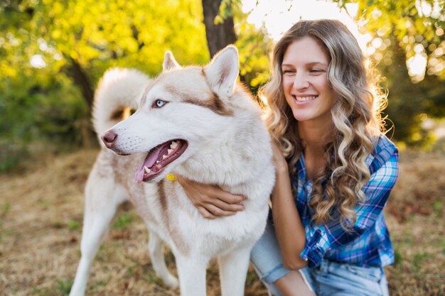 Sunny young stylish pretty smiling happy blond woman playing with dog husky breed in park on sunny summer day