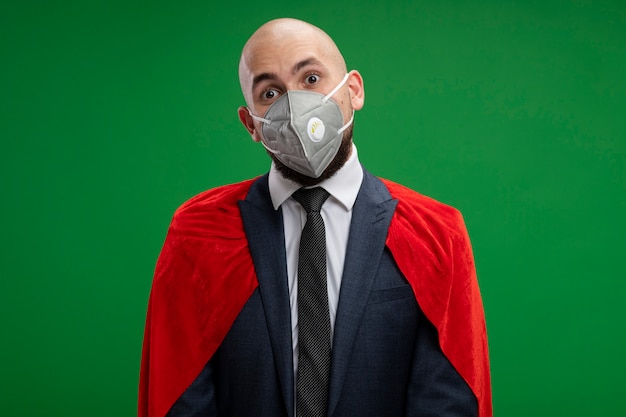 Free photo super hero bearded business man in red cape and protective facial mask  confused standing over green wall