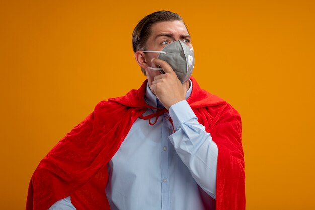 Super hero businessman in protective facial mask and red cape looking aside with pensive expression on face with hand on chin thinking standing over orange background