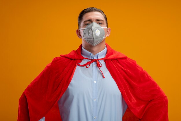 Super hero businessman in protective facial mask and red cape looking confident with arms at hip standing over orange wall