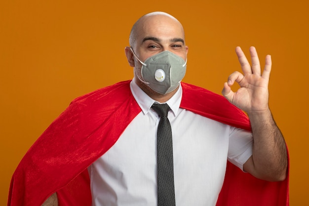 Free Photo super hero businessman in protective facial mask and red cape looking at front smiling showing ok sign standing over orange wall