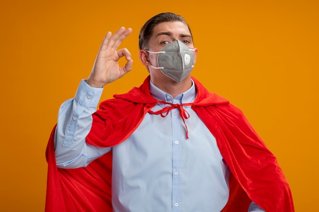 Super hero businessman in protective facial mask and red cape  smiling happy and positive showing ok sign standing over orange wall