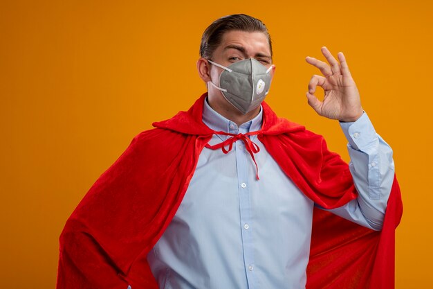 Super hero businessman in protective facial mask and red cape  smiling showing ok sign standing over orange wall