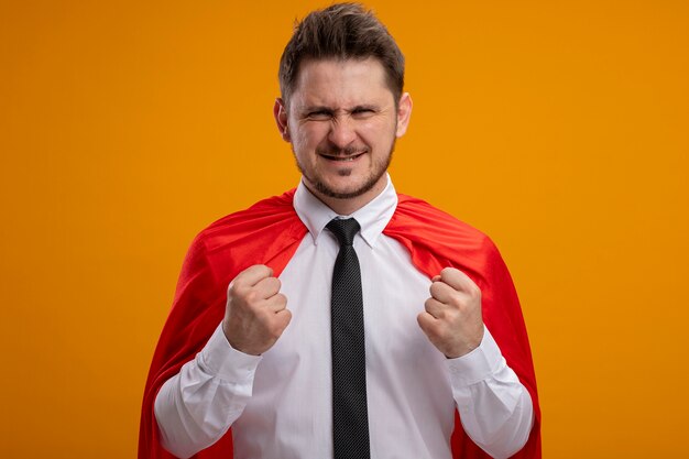 Super hero businessman in red cape clenching fists being displeased standing over orange wall