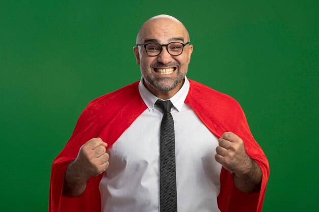 Super hero businessman in red cape and glasses clenching fist happy and excited standing over green wall