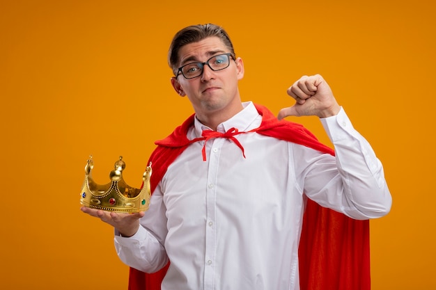 Super hero businessman in red cape and glasses holding crown pointing at himself smiling confident standing over orange wall