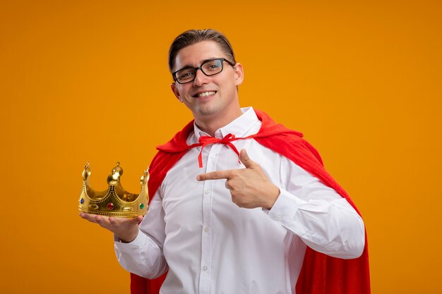 Super hero businessman in red cape and glasses holding crown pointing with index finger at it smiling confident standing over orange background