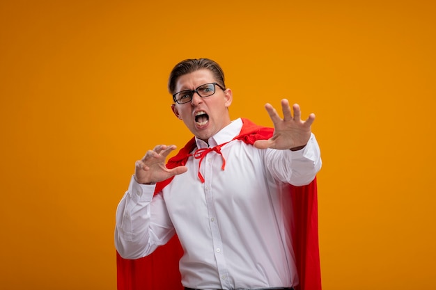 Super hero businessman in red cape and glasses looking at camera with arms out shouting with aggressive expression making defense gesture standing over orange background