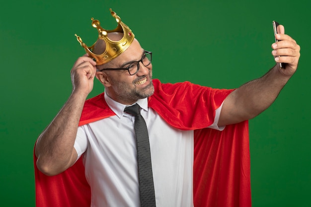 Super hero businessman in red cape and glasses wearing crown doing selfie using smartphone smiling 