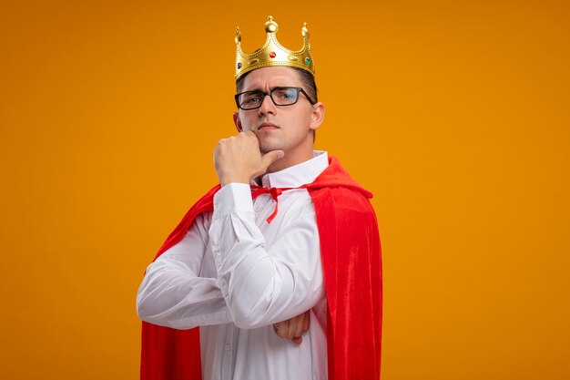 Super hero businessman in red cape and glasses wearing crown looking at camera with hand on chin with confident serious expression standing over orange background