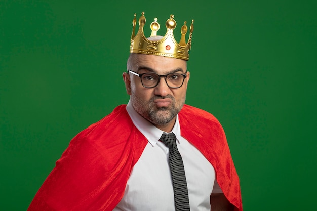 Free photo super hero businessman in red cape and glasses wearing crown looking at front self-satisfied standing over green wall