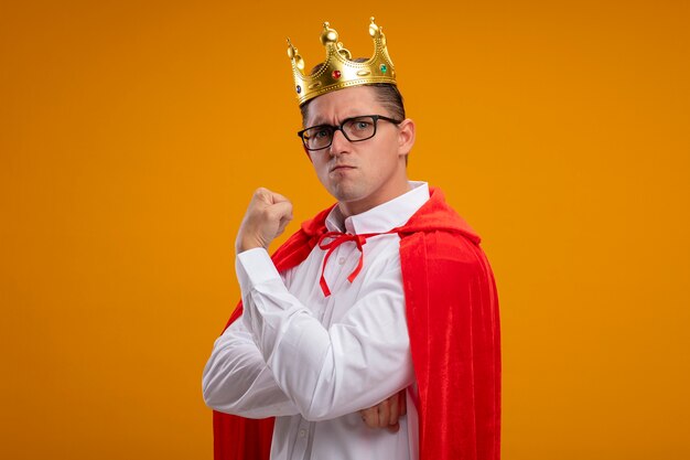 Super hero businessman in red cape and glasses wearing crown  with serious frowning face clenching fist showing strength standing over orange wall