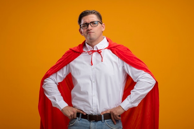 Free photo super hero businessman in red cape and glasses  with serious face with arms at hip standing over orange wall