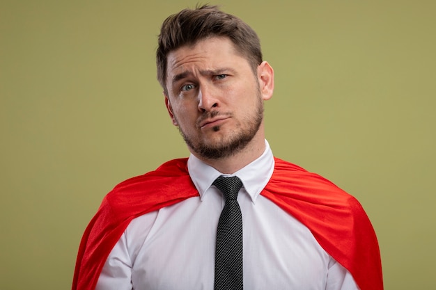 Super hero businessman in red cape lookign at camera with sad expression on face standing over green background