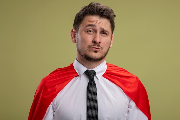 Free photo super hero businessman in red cape looking at camera being confused with sad expression standing over green background